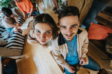Obraz premium Two high school students look up at the camera while sitting together in a relaxed indoor study setting. A casual study group scene with friends chatting and working in a living room environment.