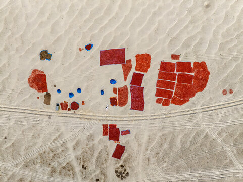 Aerial view of red chili peppers drying in rectangular patches on a sandy riverbed with workers and blue umbrellas in Bogura, Rajshahi Division, Bangladesh.