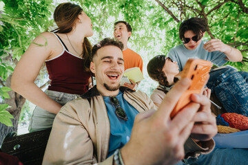 Fototapeta na wymiar High school students laugh and share a smartphone while sitting under shady trees. A group of teenagers relax together outdoors, studying and socializing in a park-like setting.