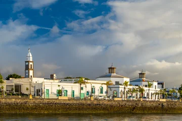 Arrecife Lanzarote waterfront view featuring historical buildings and people leisurely walking along the promenade. The serene atmosphere and clear sky create a picturesque scene © Kotangens
