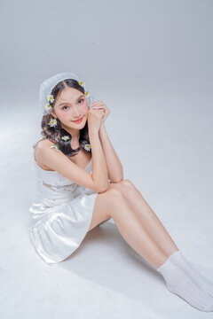 Elegant young Asian woman crouching in white satin dress with daisy flowers in hair, fashion beauty portrait hugging knees on grey background, spring bridal editorial studio shot, copy space