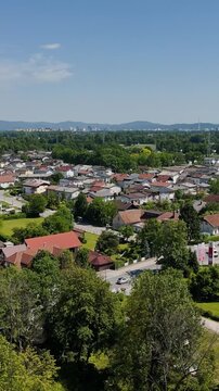 Aerial view over Tacen, Ljubljana, showing red-roof homes, green trees, distant city skyline and forested hills under a clear blue sky.