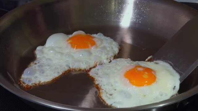 Fried eggs stick to a stainless steel pan while cooking. Spatula tries to unstick eggs from stainless steel cooking surface. Sunnyside up eggs overcook in pan while cook tries to unstick them.
