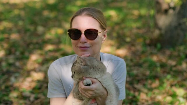 Woman holds cat gently while sitting under trees shade. She hugs her pet tenderly. Cat looks calm, eyes half-closed in trust. Sunlight filters through leaves, soft and warm.