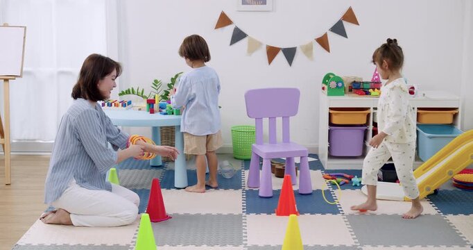 Asian female preschool teacher claps praise while preschool girl balances on chair, preschool boy stands near table, indoor daycare playroom with slide, cone marker, soft mat, motor skill training