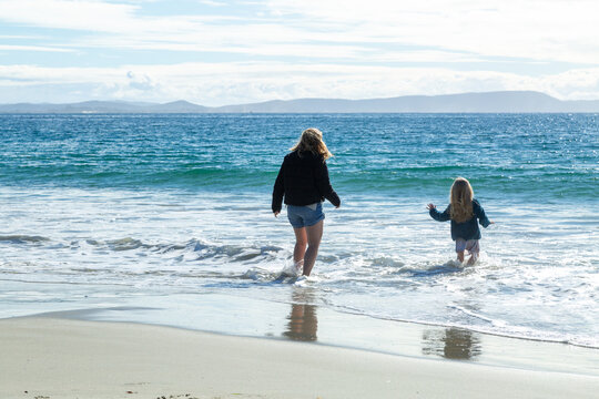 A blond mother and daughter wade into the ocean in winter clothes.