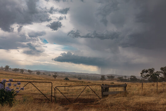 Dramatic storm clouds gather over a dry, rural Australian landscape with a weathered gate