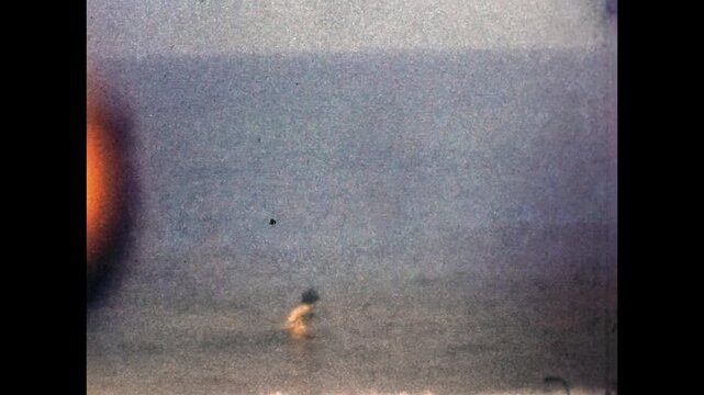 Child standing on shoreline, getting hit by waves while playing on sandy beach during nostalgic summer vacation. Family holidays memories. Super 8mm 1960s film. Old Archival retro. Vintage archive