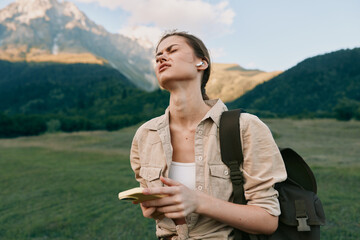 Obraz premium Young woman traveler in a green valley, notebook in hand, backpack over her shoulder, gazing toward distant mountains. Natural light highlights outdoor exploration and curiosity.