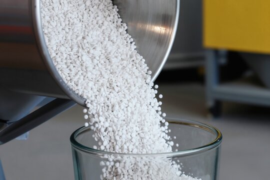 White plastic pellets pour from metal container into glass beaker. Small granules fill transparent vessel. Industrial process, material production, chemical manufacturing, factory setting.
