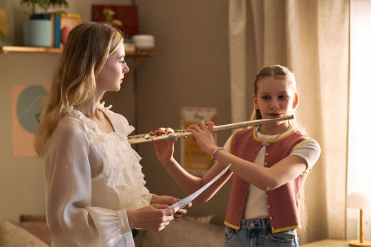 Caucasian girl playing flute while female music teacher coaching with sheet music during lesson, supporting arts education and skill development. Ideal for music school promotion