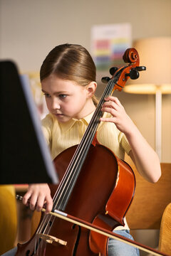 Vertical shot of Caucasian girl studying music and playing cello while reading sheet notes, showing focused arts education and skill development. Ideal for music school promotion