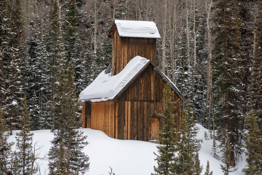 Aerial view of a historic wooden mine building covered in deep snow surrounded by a dense forest of pine and aspen trees along the Million Dollar Highway in Ouray, Colorado, United States.