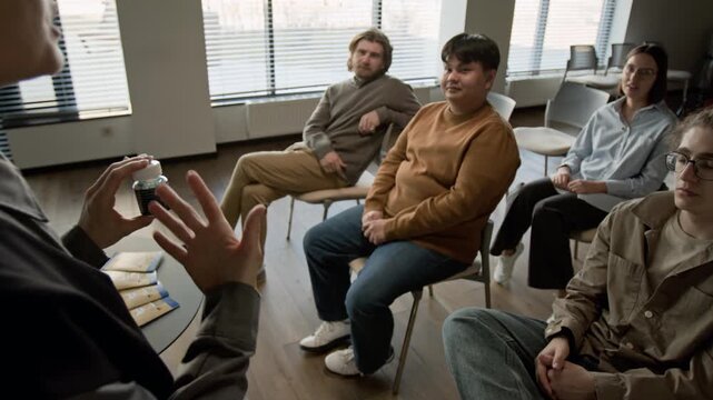Female psychotherapist holding medicine bottle, explaining treatment to diverse group of patients during therapy session. Clip for mental health and pharmaceutical ads