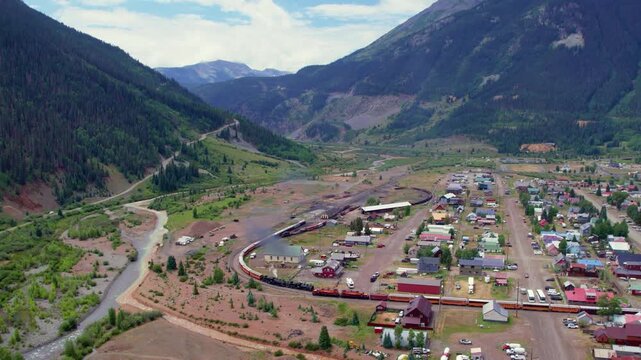 Aerial view of historic Durango to Silverton train pulling into train station at Silverton, CO. Steam coming out of engine as train goes along tracks. San Juan Mountains and Animas River in backdrop.