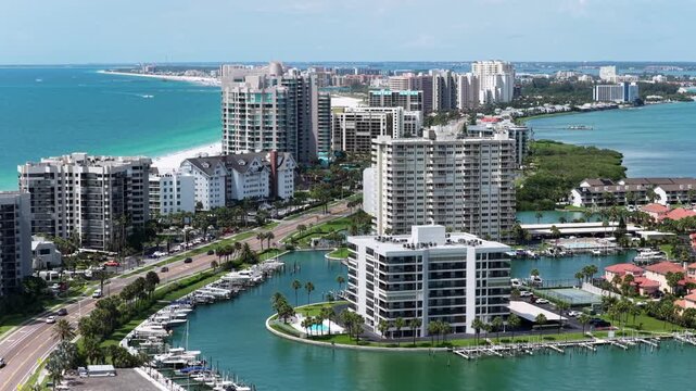 Luxury high-rise condos flank Clearwater Beach waterway and marina, with turquoise Gulf waters and white sand shoreline under blue skies in Clearwater Beach, Florida, USA. Aerial drone view.