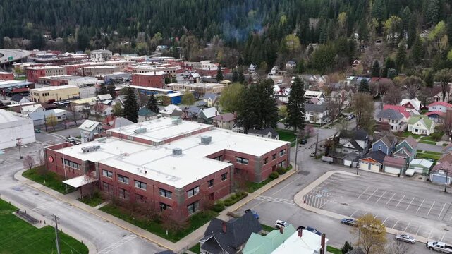 Colorful single-family homes and tree-lined streets spread across residential blocks beside historic brick commercial buildings under gray skies in Wallace, Idaho, USA. Aerial drone view.