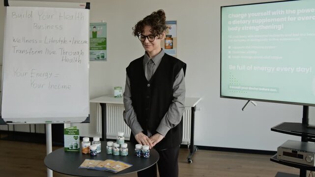 Portrait of young Caucasian woman with glasses standing in office next to flipchart and table with dietary supplements, smiling at camera. Footage for wellness business marketing