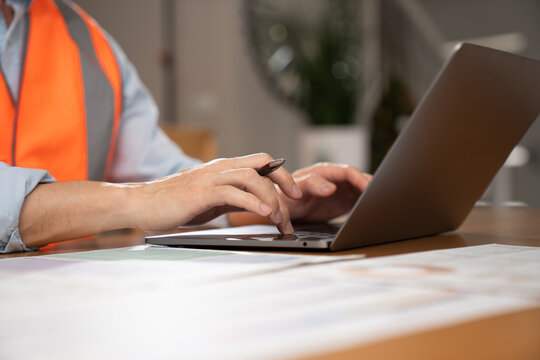 Close-up view of an engineer or safety officer wearing a high-visibility vest and working on a laptop while reviewing documents and holding a pen in a modern office environment.