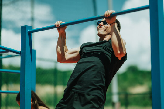 A man in sunglasses performs a pull-up on blue outdoor bars showing upper-body strength. An outdoor workout scene with calisthenics training on a sunny day.