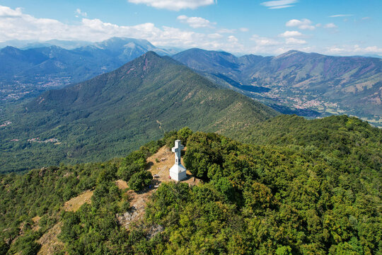 Aerial view of the white summit cross on Monte Musine surrounded by lush green forests and the Alpine mountain range under a blue sky in Caselette, Piedmont, Italy.