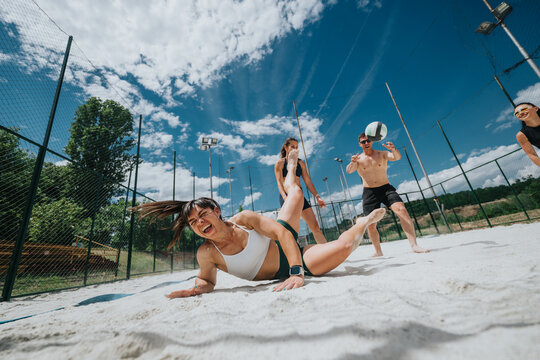 A group of young friends play beach volleyball as a woman dives in the sand to reach the ball. The outdoor game shows energetic action, laughter and summer fun on a fenced sand court.