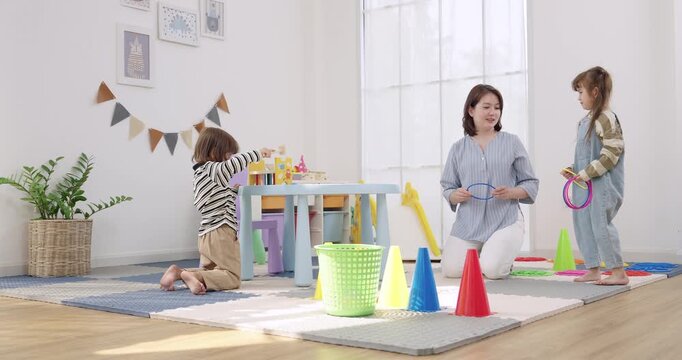 Asian female preschool teacher reaches guiding toddler boy crawling, toddler girl prepares ring toss near cone target, indoor daycare playroom, coordination training, energetic early education