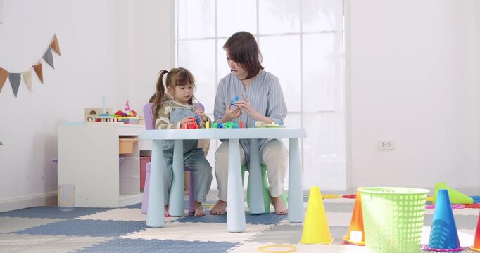 Asian female preschool teacher beside toddler girl, observing toddler boy arrange peg stack pieces on table, bright classroom lesson, patience coaching, cognitive development, counting color task.