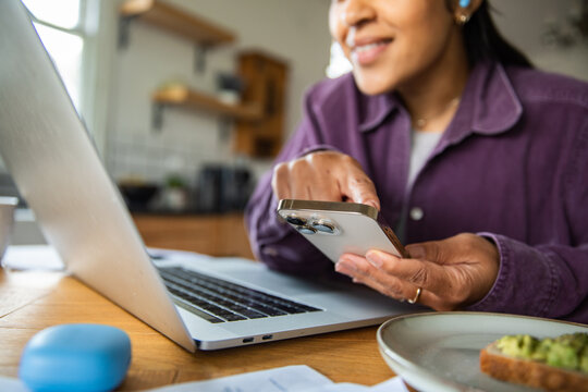Smiling woman paying bills on smartphone at home kitchen table