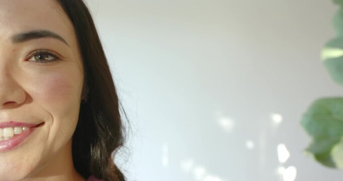 Adult woman responding to light, smiling, wearing maroon top by pale wall with plant, copy space