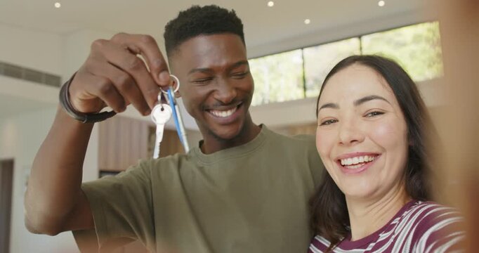 Diverse couple at home holding up keyring with blue fobs toward camera laughing, celebrating move