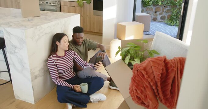 Diverse couple sitting on floor by island, unpacking boxes while checking handheld device and mug