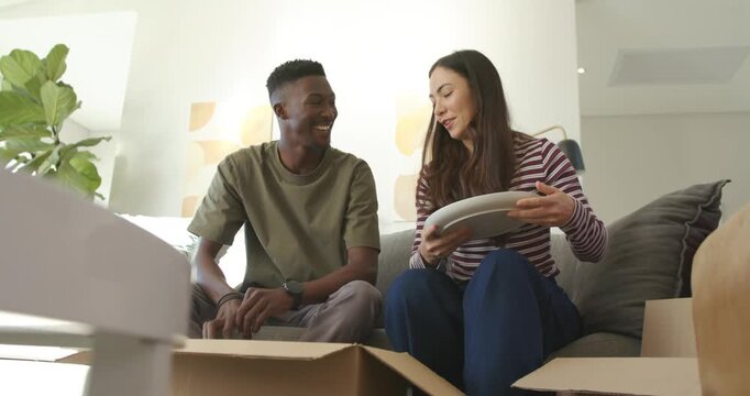 Diverse couple unpacking boxes on couch woman reaching box and handing plates while man stacking
