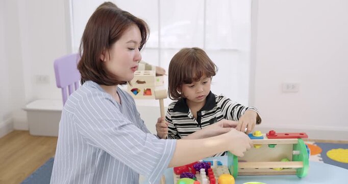 Asian female preschool teacher cheering toddler boy holding toy hammer after workbench success, daycare positive reinforcement, joyful learning, confidence building, coaching, teamwork play.