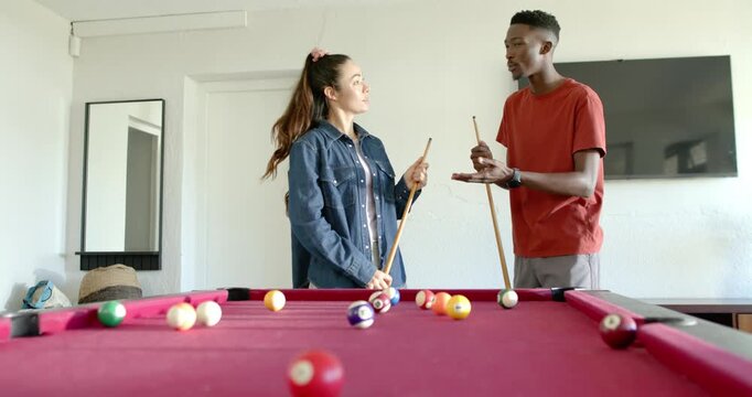 Diverse couple behind red-felt table, man cupping in red tee explaining shot holding cue near balls