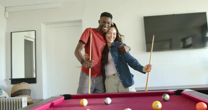 Couple holding cues behind red pool table at home, smiling to camera man reaching leaning laughing