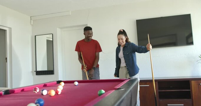 Diverse couple chatting, man stepping closer teaching her cue grip at pool table with racked balls