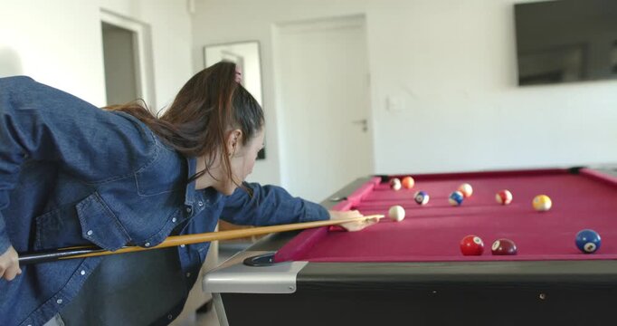 Asian woman 20s in denim jacket approaching pool table, lining cue and striking cue ball practicing