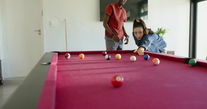 Diverse couple playing pool at home, woman lining and striking cue, sending red ball across table
