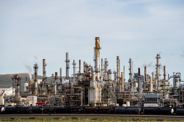 Obraz premium Industrial complex with tall towers and pipes. Cloudy sky looms over distant mountains. Metal structures dominate the arid landscape. No visible animals or plants in the scene