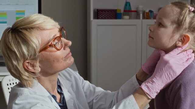 Medium closeup of Caucasian female pediatrician palpating neck of child girl during medical checkup in clinic. Footage is ideal for pediatric healthcare services and medical clinic promotions