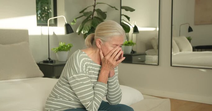 Senior female covering face with both hands on bed edge in bedroom, feeling overwhelmed