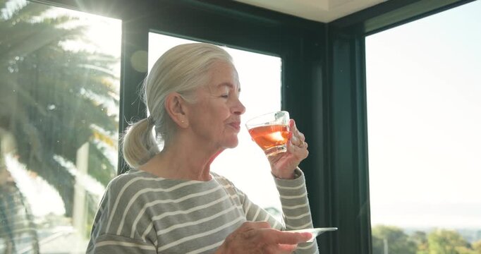 Senior woman in striped top holding glass cup, saucer in sunroom, looking out windows, sipping tea