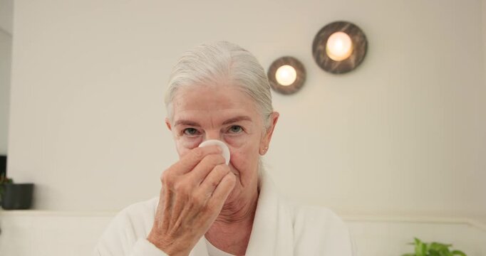 Senior woman raising cotton pad, wiping face, removing makeup at home vanity in white bathrobe