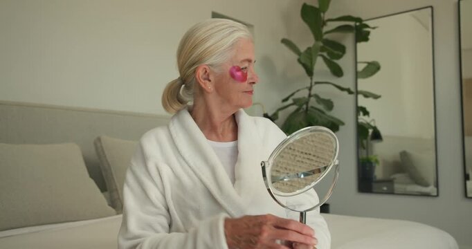 Senior woman after applying pink eye patches on bed in white bathrobe smoothing skin with mirror