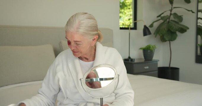 Senior woman on bed in white robe looking into mirror applying pink eye patches for skincare