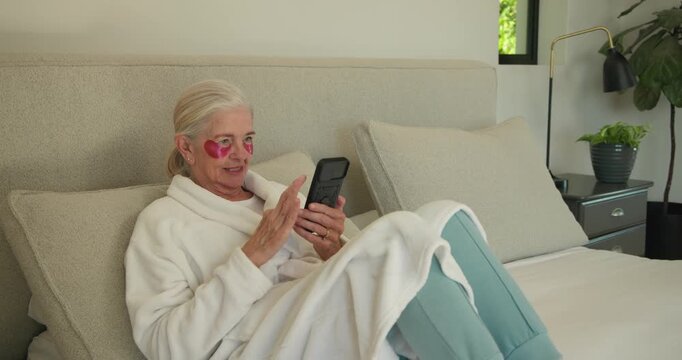 Senior woman in white robe on bed with pink patches, looking at phone, scrolling and smiling