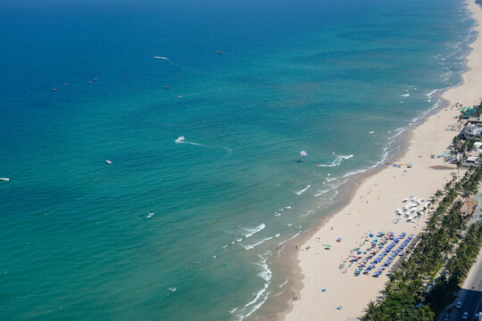 Many people swim in the blue ocean and relax on the sandy beach under sun umbrellas. A parasailer glides across the water near the shore on a bright, sunny day