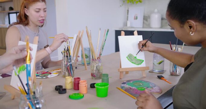 Diverse female friends painting on easels at kitchen table, third joining to create leaf paintings