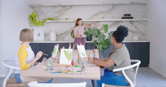 Diverse female painters watching redhead teacher checking plant and teaching leaf shapes on canvas
